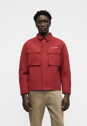 Man wearing a red Columbia jacket with two large front pockets, beige pants, standing against a plain white background.