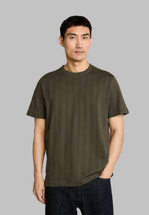 Young man with short black hair wearing an olive green short-sleeve shirt and dark pants, standing against a gray background.