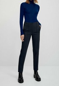 Vrouw in een strak navyblauw turtleneck, zwarte tailored broek met voornaad, handen in de zakken, en zwarte enkellaarsjes op een witte achtergrond.