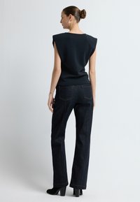 Top senza maniche nero con spalle strutturate, abbinato a jeans a vita alta blu scuro con cuciture arancioni e tasche posteriori. Tacchi neri.