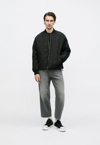 Νέος άνδρας ντυμένος με μαύρο καπιτονέ bomber μπουφάν, ξεθωριασμένο γκρι cropped τζιν, λευκές κάλτσες και μαύρα sneakers χαμηλού προφίλ, στέκεται μπροστά σε απλό φόντο.