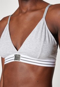 Bralette gris avec un design en V profond, fabriqué en tissu doux, mettant en avant une bande blanche avec des rayures noires et une étiquette de marque.