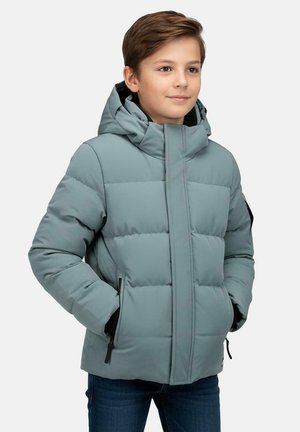 Garçon aux cheveux courts et bruns portant une veste matelassée bleu clair avec capuche et les mains dans les poches, debout devant un fond uni.