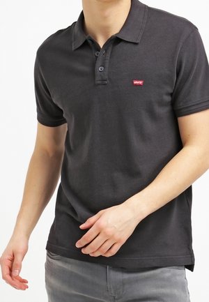 Mann trägt ein schwarzes Levi's-Poloshirt mit kurzen Ärmeln und zwei Knöpfen, kombiniert mit hellgrauen Hosen, gezeigt von den Schultern bis zur Hüfte.