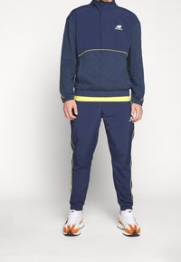 Námořnicky modrá bundička s žlutými akcenty, zipem u límce a elastickými manžetami, doplněná o sladěné námořnicky modré kalhoty s bočními pruhy a přiléhavým střihem.