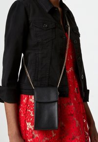 Musta nahkainen olkalaukku, jossa on läppätasku ja kultainen ketjuhihna, päällä punaisen pitsimekon ja istuvan mustan farkkutakin kanssa.