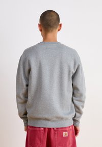 Felpa grigia con collo rotondo e polsini a coste, indossata su pantaloni rossi. Il tessuto sembra morbido, con una vestibilità comoda e senza grafiche visibili.