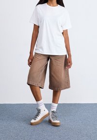 Bílý bavlněný tričko, hnědé džínové culoty a tenisky s vyšším střihem, vyrobené z texturované bílé látky s vzorovanými tkaničkami, nošené se pruhovanými ponožkami.