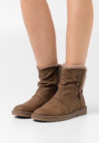 Botas tobilleras marrones con un puño de piel sintética, cierre de cremallera y dos botones decorativos. Fabricadas con material suave, cuentan con una suela plana y una superficie texturizada.