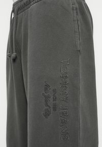 Detailný záber na tmavosivé teplákové nohavice s elastickým pásom a vyšitým logom Tommy Jeans na stehne.