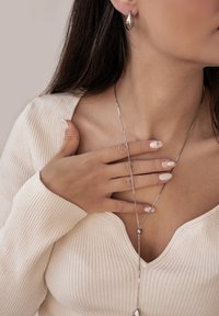 Zilveren ketting met een druppelvormige hanger, gedragen over een crèmekleurig ribgebreid topje. Close-up van handen met lichtroze nagels, één hand leunend op de sleutelbeen.