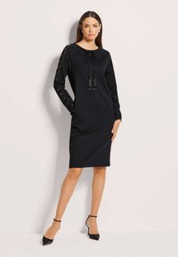 Frau in einem schwarzen knielangen Kleid mit Spitzenteilern und vorderem Knoten, die mit einer Hand in der Tasche steht und schwarze High Heels trägt.