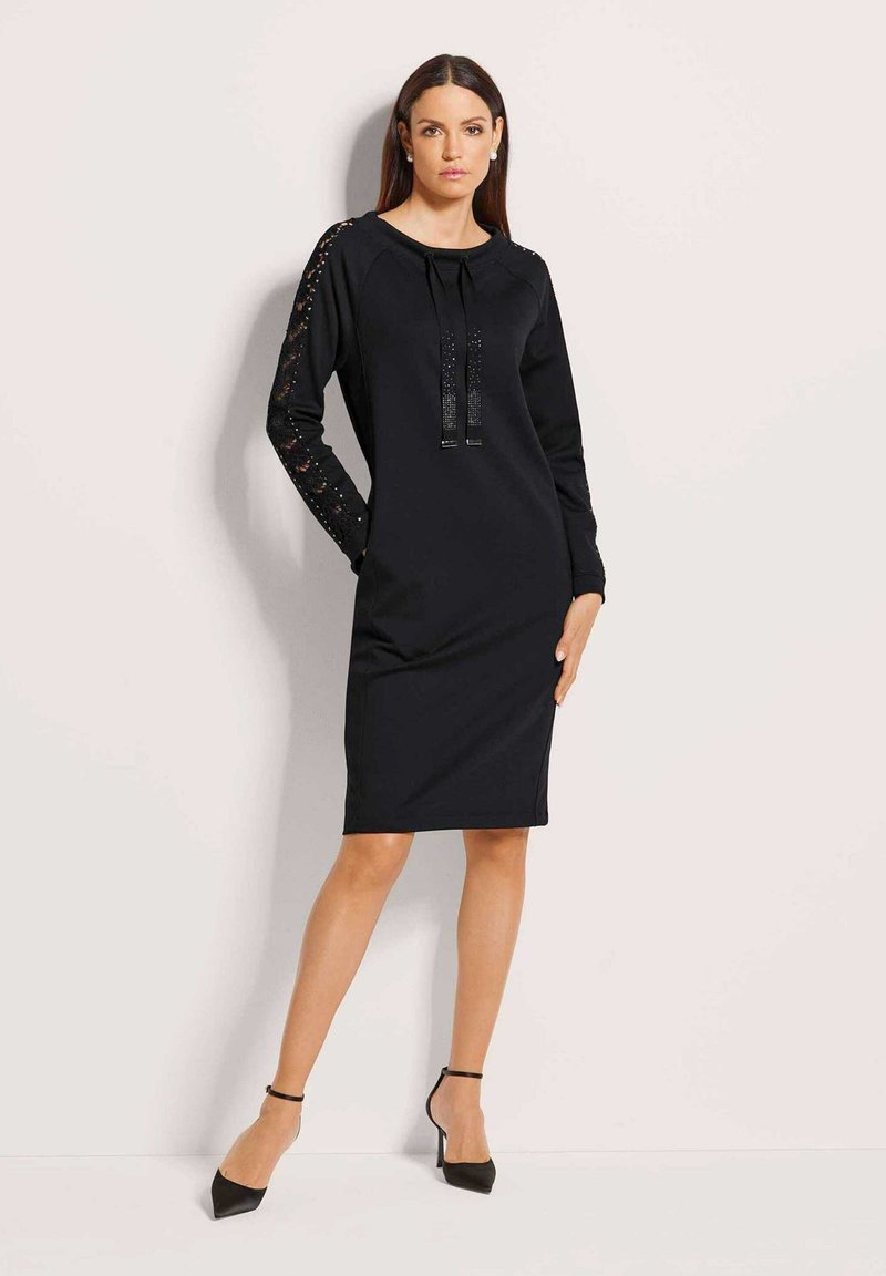 Frau in einem schwarzen knielangen Kleid mit Spitzenteilern und vorderem Knoten, die mit einer Hand in der Tasche steht und schwarze High Heels trägt.