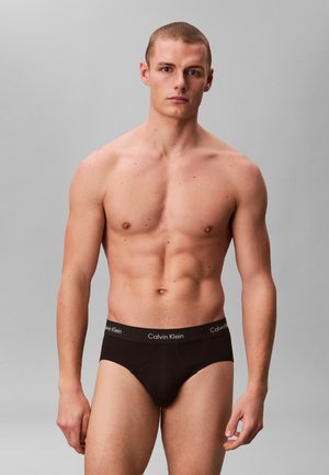 Sprawny młody mężczyzna z krótkimi włosami, ubrany w czarne slipy Calvin Klein, stoi na jednolitym szarym tle.