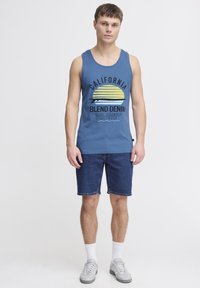 Top de bumbac albastru fără mâneci, cu un imprimeu grafic, asortat cu șorturi din denim albastre. Modelul poartă adidași gri și șosete albe. Design simplu, croială lejeră.