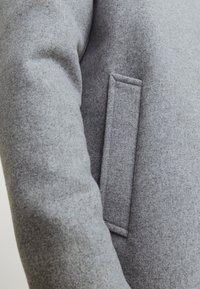 Abrigo de lana gris con un diseño elegante, textura suave y un bolsillo lateral con un sutil detalle de costura. La tela parece cálida y duradera.