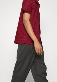 Hombre con una camisa roja de patrón de manga corta y pantalones oscuros grises, mostrado desde el cuello hasta la mitad del muslo, caminando hacia la izquierda.