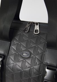 Sac synthétique matelassé noir avec des motifs géométriques, deux zips en métal, et un badge logo Lacoste. Bretelles ajustables avec des accents en métal.