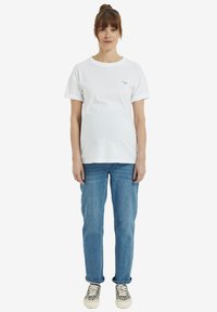 Junge Frau mit braunen Haaren zu einem Dutt gebunden, trägt ein weißes T-Shirt mit hochgerollten Ärmeln, blaue Jeans und Turnschuhe mit Leopardenmuster, steht vor einem weißen Hintergrund.