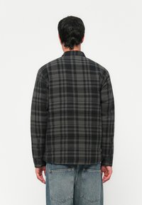 Man facing away wearing a black and gray plaid long-sleeve shirt and light blue denim jeans with visible back pockets.