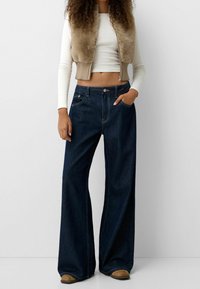 Hoch taillierte, weit geschnittene Jeans aus dunklem Denim mit vorderen Taschen. Kombiniert mit einer cropped beigen Fleece-Weste und einem enganliegenden weißen Langarmshirt.