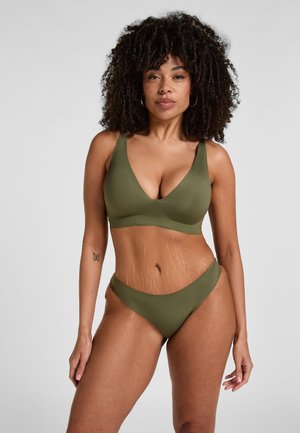 Frau mit lockigem Haar, die einen olivgrünen BH und Slip trägt, vor einem schlichten hellgrauen Hintergrund stehend, mit einem Schmetterlingstattoo am Arm.