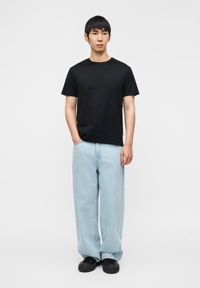 T-shirt noir en coton à manches courtes, coupe décontractée, associé à un jean droit bleu clair et des chaussures noires, posé devant un fond blanc.