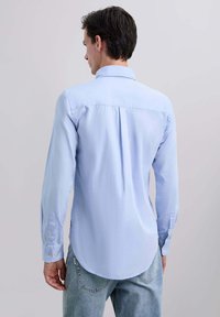 Chemise à manches longues bleu clair avec un col classique, un plissé au dos et des poignets à boutons, fabriquée dans un tissu lisse. Associée à un jean en denim délavé.