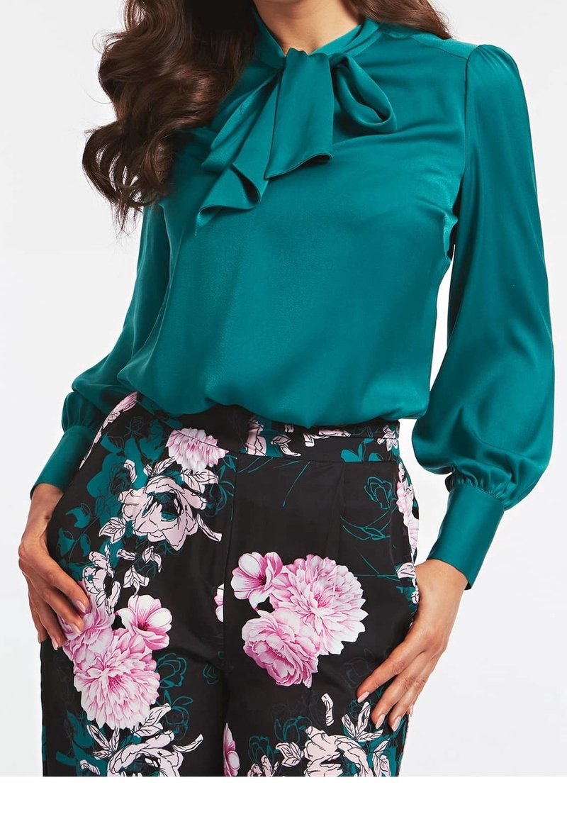 Tealfärgad satinblus med en slagkrage och rosett, puffärmar, bärs tillsammans med svarta blommiga byxor som har rosa pioner och gröna accenter.