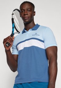 Blå polo shirt med krage, med ljusblå och vita ränder över bröstet. Bärs av en person som håller en tennisracket.