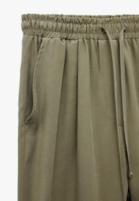 Olivenfarvede bukser med elastisk talje, snøre og to sidelommer. Stoffet har en glat tekstur med en let glans.