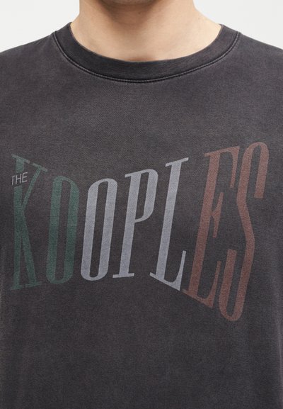 Άτομο που φοράει σκούρο γκρι μπλουζάκι με στρογγυλή λαιμόκοψη και μεγάλο, πολύχρωμο κείμενο "THE KOOPLES" στο στήθος.