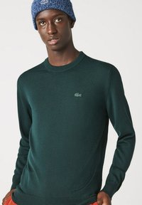 Maglione lavorato a maglia verde scuro con colletto rotondo, con un logo ricamato discreto sul petto. Il tessuto appare liscio ed elastico.