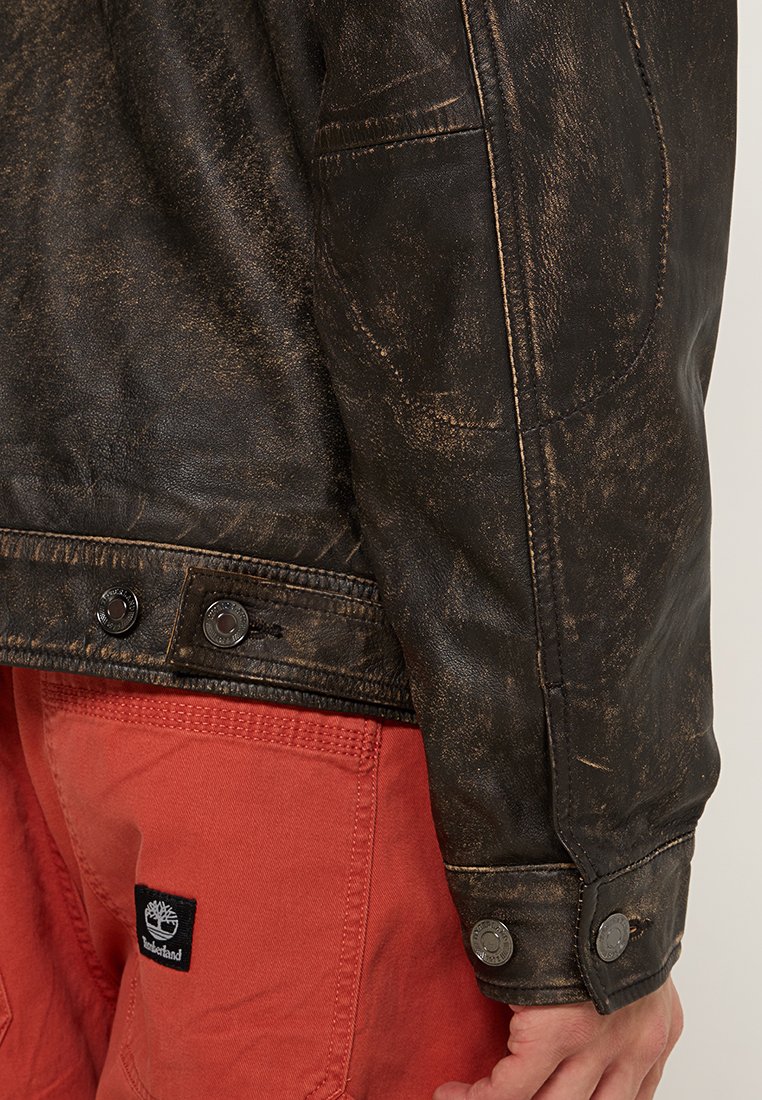 Czarna skórzana kurtka z efektem postarzenia, z mosiężnymi akcentami i zatrzaskami, w połączeniu z czerwonymi spodniami z naszywką logo na kieszeni.