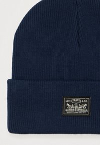 Navy knitted beanie with ribbed texture and turned-up cuff. Features a rectangular black label with white text and graphic detailing.