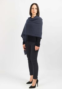 Un avvolgente in lana blu navy si drape sulle spalle, abbinato a un maglione nero e pantaloni aderenti. La modella indossa scarpe con tacchi alti nere.