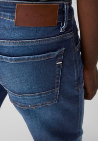 Jeans de mezclilla en azul oscuro con una textura desgastada, con un parche de cuero marrón de Marc O'Polo, bolsillo trasero visible y costuras en blanco.