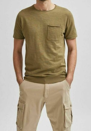 Homme portant une chemise vert olive à manches courtes avec poche poitrine et un pantalon cargo beige, les mains dans les poches.