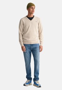 Beiger V-Ausschnitt-Pullover aus weichem Material, kombiniert mit hellblauen Slim-Fit-Jeans und sportlichen Sneakers mit blauen Akzenten. Grüner Strickmütze auf dem Kopf.