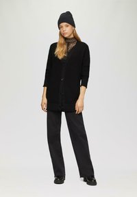 Zwarte gebreide cardigan met v-hals en knoopsluiting, gecombineerd met zwarte wijde pantalons en een bijpassende gebreide beanie. Textuurdetails zichtbaar.