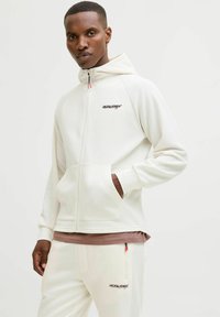 Bela zip-up kapuca s kapuco na vezico, rebrastimi stranskimi paneli in sprednjimi žepi. Združena z usklajenimi trenirkami z logotipnimi podrobnostmi.