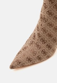 Chaussure en tissu marron à bout pointu avec motif monogramme marron foncé répété sur fond beige texturé.