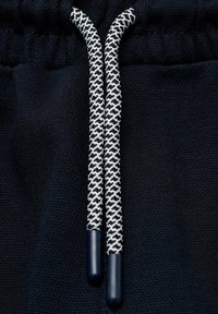 Černá látka s texturovaným povrchem má bílé a černé tkané šňůrky, které končí na tmavých plastových špičkách, což zdůrazňuje kontrast barev a designu.