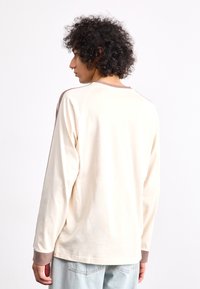 Μακρυμάνικη μπλούζα από ελαφρύ μπεζ βαμβάκι, με καφέ λεπτομέρειες στους ώμους και στις μανσέτες. Λείος υφιστάμενος και σχέδιο χαλαρού εφαρμογής.