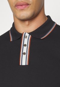 Gros plan d'une personne portant un polo noir avec un col et une patte rayée blanche, marron et bleu marine avec trois boutons.
