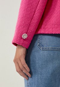 Giacca fucsia con texture e un bottone decorato con strass trasparenti, indossata sopra un denim blu con tasca a taglio diritto.