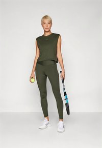 Olívazöld sportos öltözék, ujjatlan felsővel és testhezálló leggingsszel. A szövet sima tapintású, fehér sportcipővel és egy teniszlabdával.