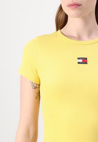 T-shirt jaune à côtes avec manches courtes, arborant un label "TOMMY JEANS" noir, rouge et blanc sur la poitrine. Texture de tissu lisse.