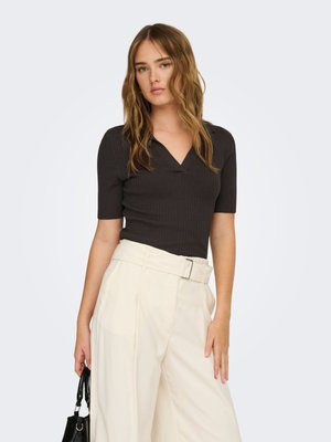 Kvinne med bølgete hår iført en svart V-hals kortermet topp og høyhalsede vide kremfargede bukser, holder en svart veske ved siden av seg.