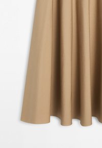 Beige blødt stofgardin hængende med bløde folder og en glat tekstur mod en hvid baggrund.